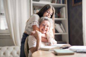 Young woman embraces an older woman from behind at a desk with books, smiling warmly in a cozy study.