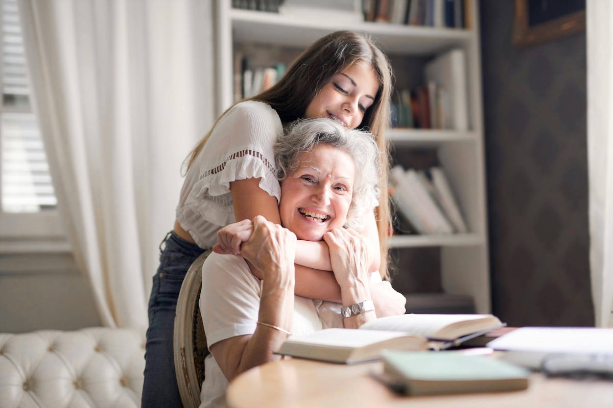Young woman embraces an older woman from behind at a desk with books, smiling warmly in a cozy study.