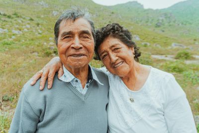 Smiling elderly couple with arms around each other outdoors in a grassy, hilly landscape.