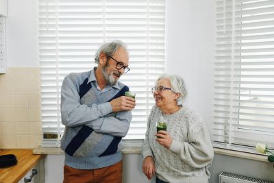 Two elderly people sharing a chat over coffee mugs in a bright kitchen by the window blinds.