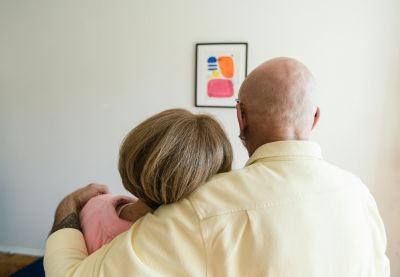 Two people standing with their arms around each other, seen from behind, in a room with a framed artwork on the wall in the background.