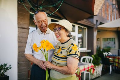 An elderly couple stands outside a cafe, smiling as the man offers a bouquet of yellow flowers to the woman.