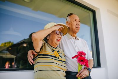 Older couple standing by a window; the woman shields her eyes from the sun with a hat, while the man holds a pink bouquet.