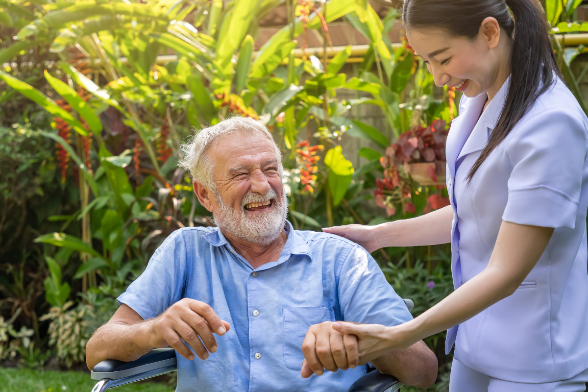 Caregiver in a lavender uniform gently helps an elderly man in a wheelchair outdoors, both smiling in a sunny garden.
